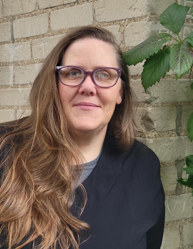 A woman with long brown hair and glasses stands in front of a brick wall, wearing a black top. There is a green plant with large leaves nearby.