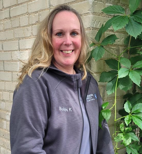 A woman with long blonde hair wearing a gray jacket embroidered with "Bobbi K" stands smiling beside a brick wall and green leafy vine.