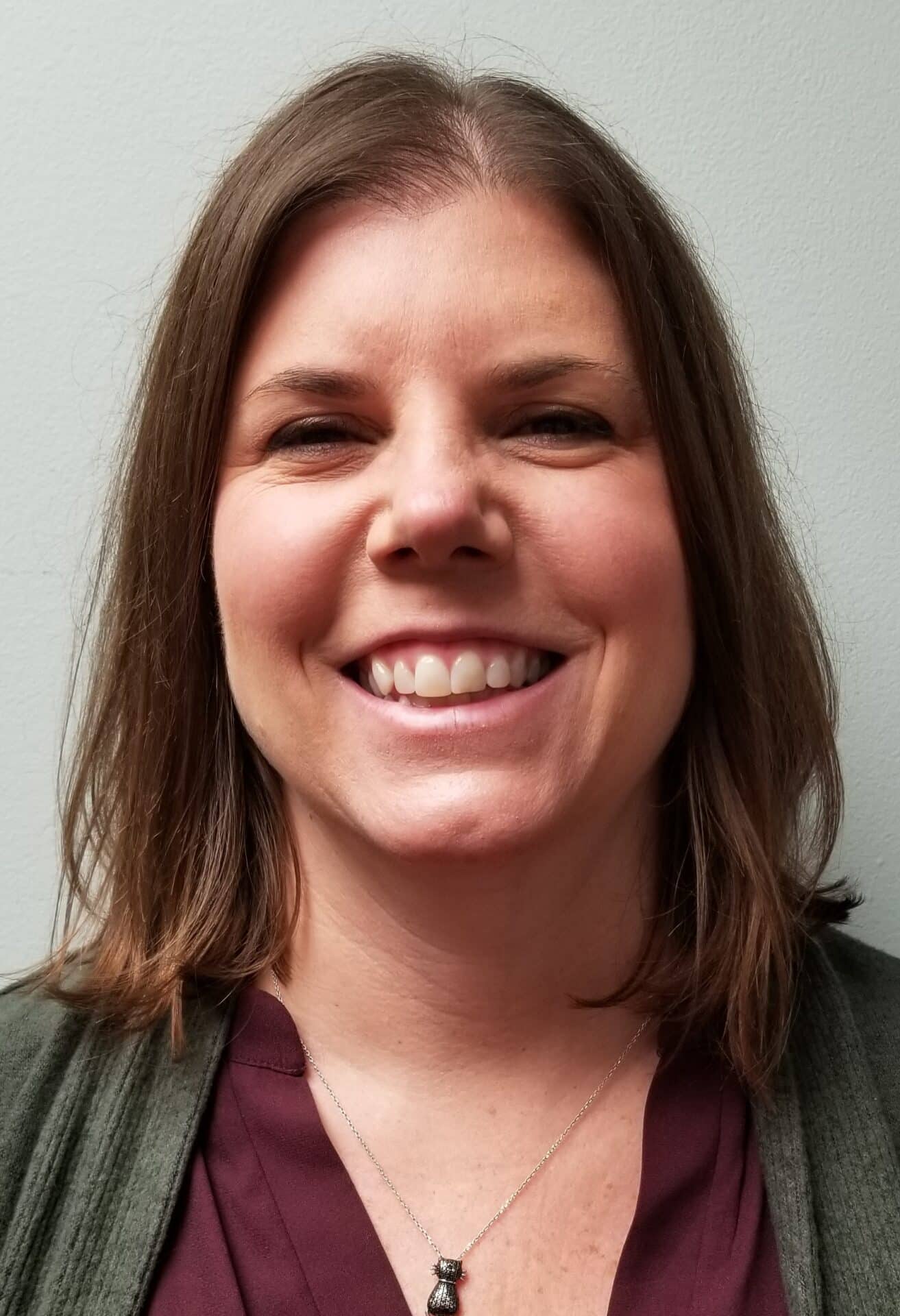 A person with shoulder-length brown hair is smiling while facing the camera. They are wearing a dark top and a necklace.