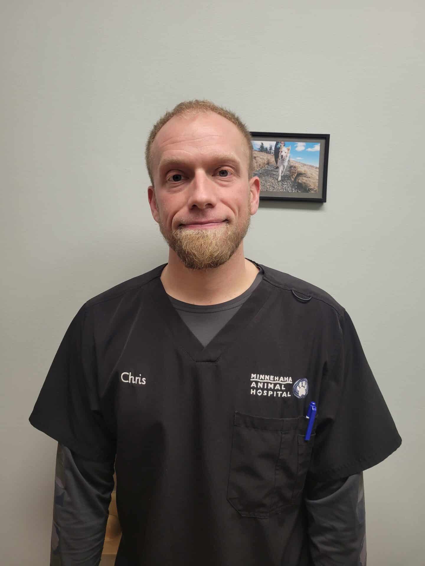 A man in a black veterinary uniform with "Chris" on the chest stands in front of a wall with a small framed picture.