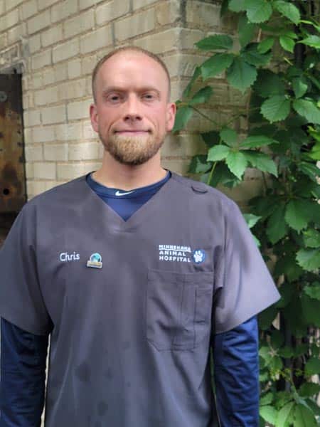 A man in gray scrubs with a name tag reading "Chris" stands in front of a brick wall and green leafy plant. His shirt says "Minnesota Animal Hospital.