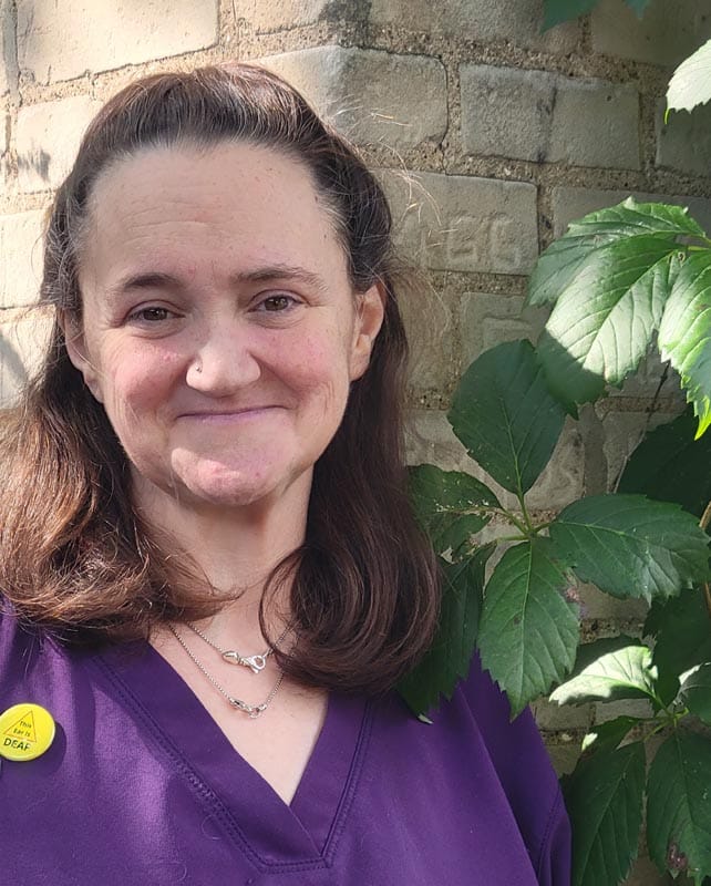 A woman with long brown hair, wearing a purple top and a yellow "I am deaf" pin, stands in front of a brick wall with green leafy plants beside her.