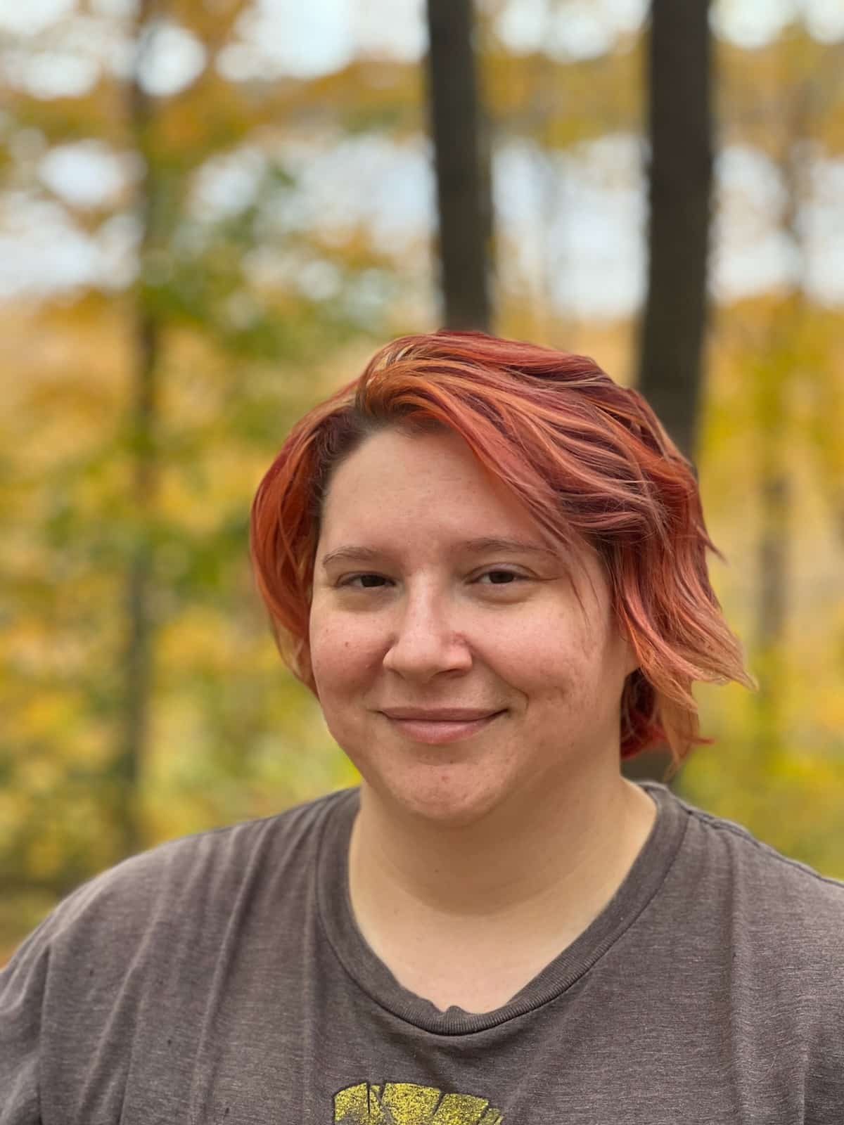 Person with short, red hair smiles in a wooded area with autumn leaves in the background.