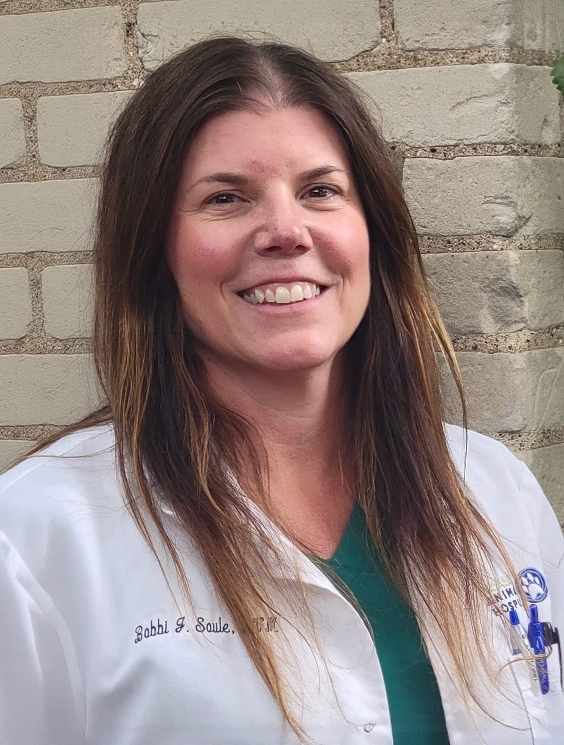 A woman with long brown hair wearing a white lab coat stands in front of a light-colored brick wall, smiling at the camera.