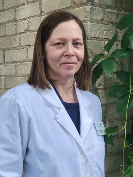 A woman wearing a white lab coat and name badge stands in front of a brick wall with green leaves nearby.