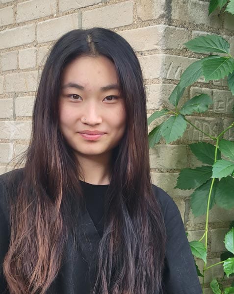 A woman with long dark hair and a nose piercing stands in front of a brick wall with green leafy plants nearby.