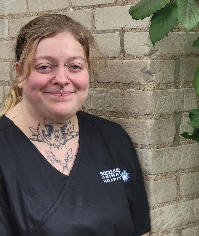 A person wearing black scrubs with a "Minnehaha Animal Hospital" logo stands in front of a brick wall, smiling.