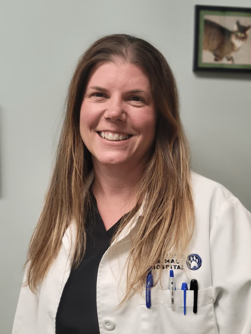 Smiling woman wearing a white lab coat with pens in the pocket, standing indoors. There's a framed animal photo on the wall behind her.