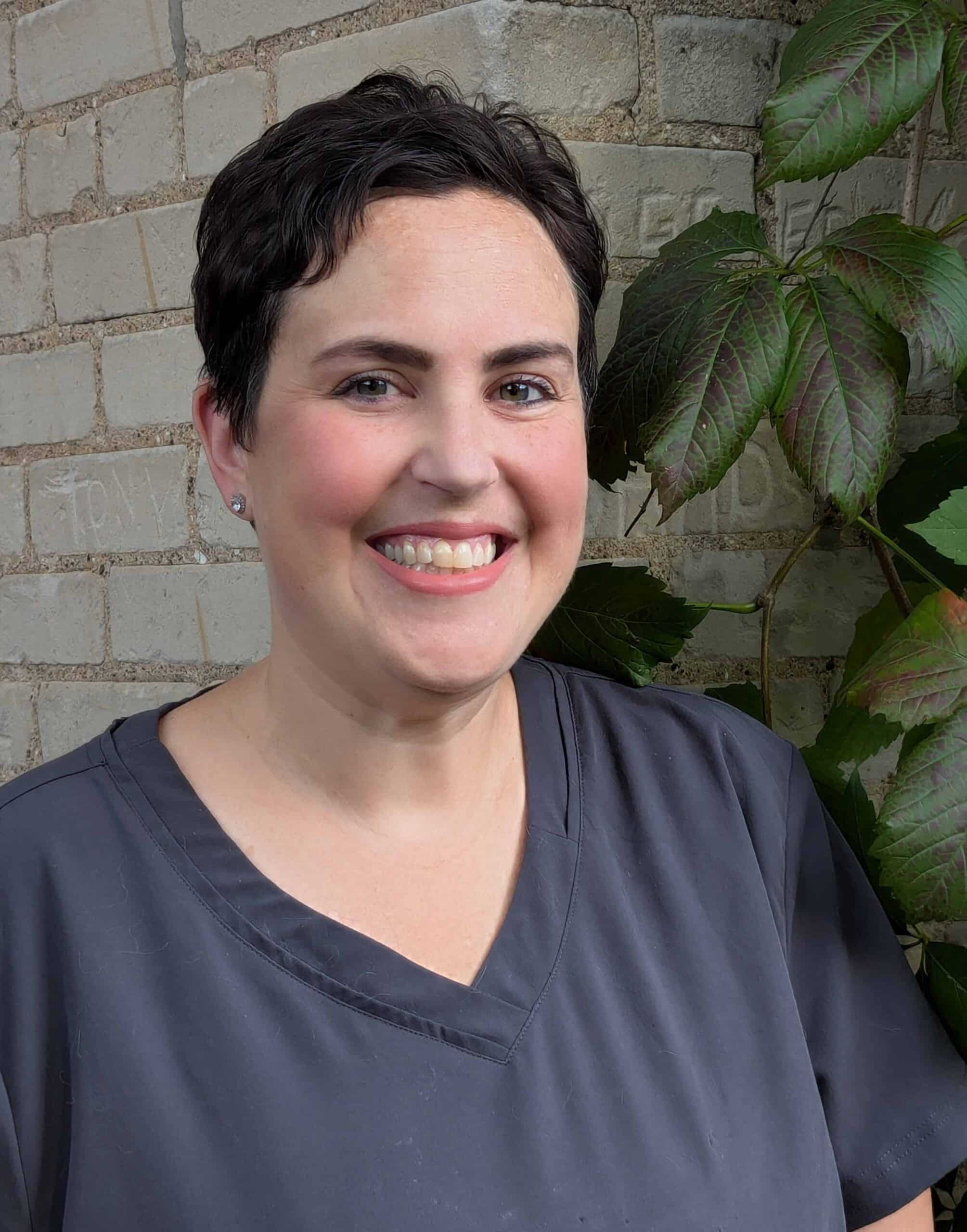 A person with short dark hair, wearing a dark gray scrub top, smiles while standing in front of a brick wall with green leaves in the background.