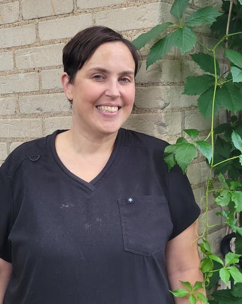 Person with short dark hair wearing a black shirt stands in front of a brick wall with green leafy vines growing along it, smiling at the camera.