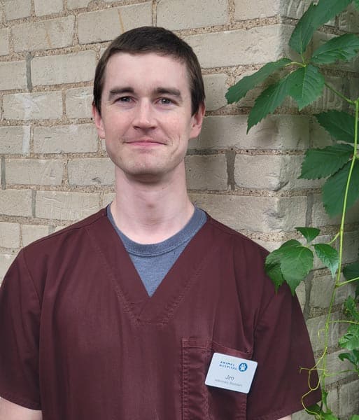 A person wearing maroon scrubs and a name badge stands in front of a brick wall with green leafy vines.