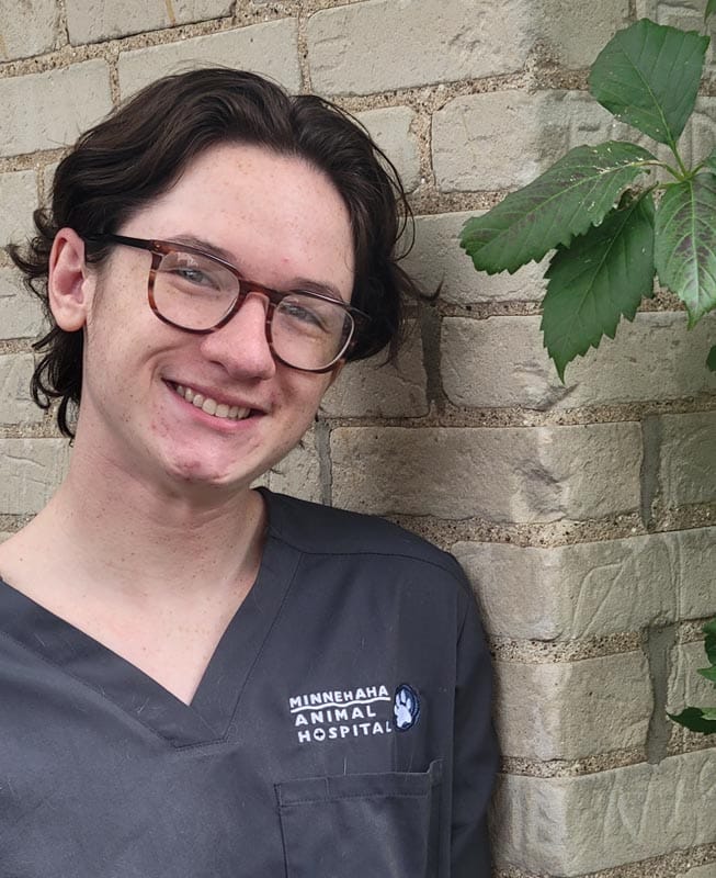 Person with glasses wearing gray scrubs labeled "Minnehaha Animal Hospital" stands and smiles in front of a brick wall with green leaves visible on the right.