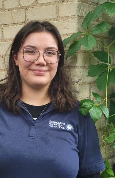 A person wearing glasses and a navy "Minnehaha Animal Hospital" shirt stands in front of a brick wall with green vines.
