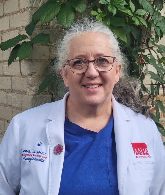 A woman with gray hair and glasses wearing a white lab coat and blue scrubs stands in front of a brick wall with green leaves in the background.