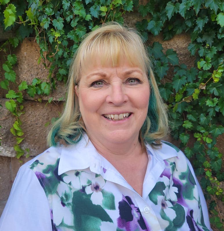 A woman with blonde hair in a floral shirt stands outdoors in front of a wall covered in green ivy.