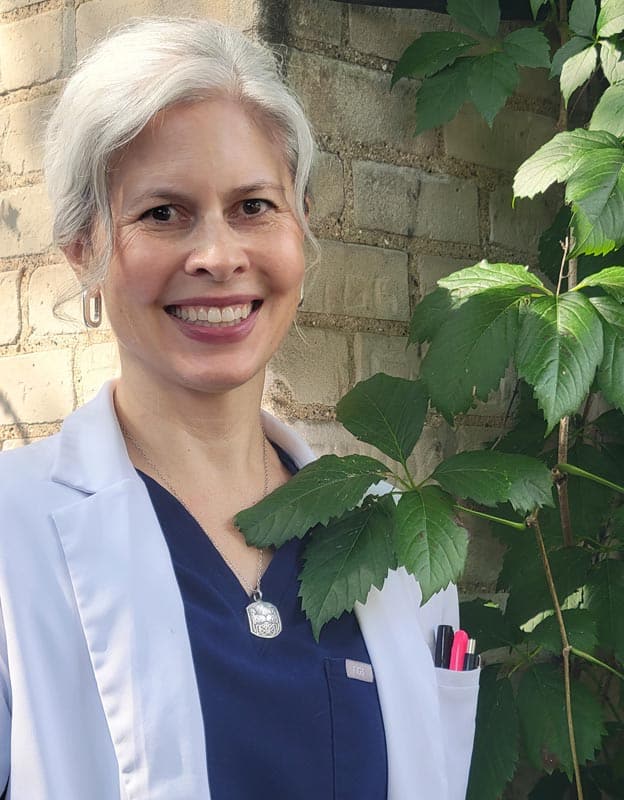 A person with gray hair wearing a white lab coat and navy blue scrubs stands outside near a leafy plant, smiling at the camera. Pens are visible in the coat pocket.